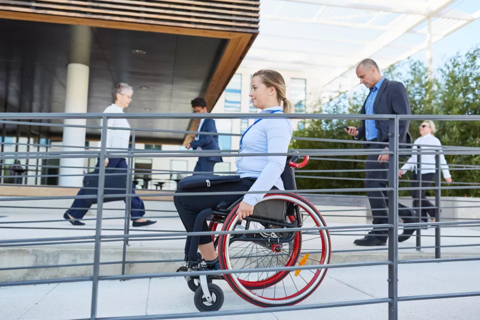 Woman in wheelchair using outdoor accessibility ramp with people walking in background